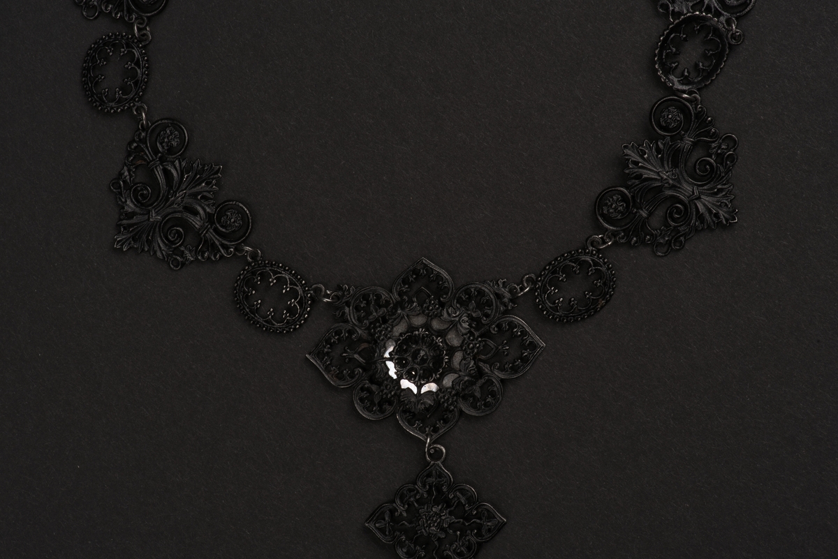 Svart halsband av gjutjärn.
Halsbandet består av omväxlande större och mindre utsirade länkar i nygotisk stil, som är sammanfästa med runda små länkar. Mitt fram hänger ett större likformat kors, som är något skadat. Spännet är utformat som två händer.