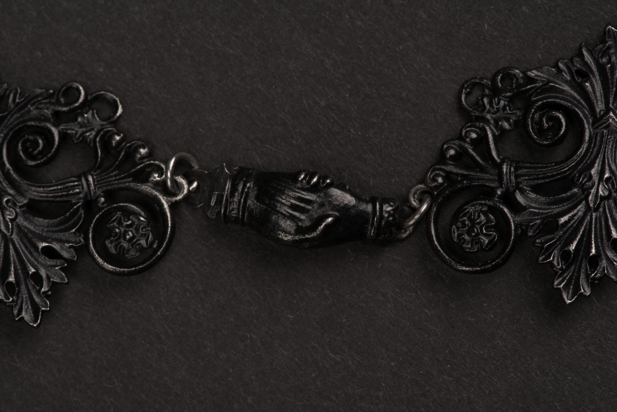 Svart halsband av gjutjärn.
Halsbandet består av omväxlande större och mindre utsirade länkar i nygotisk stil, som är sammanfästa med runda små länkar. Mitt fram hänger ett större likformat kors, som är något skadat. Spännet är utformat som två händer.