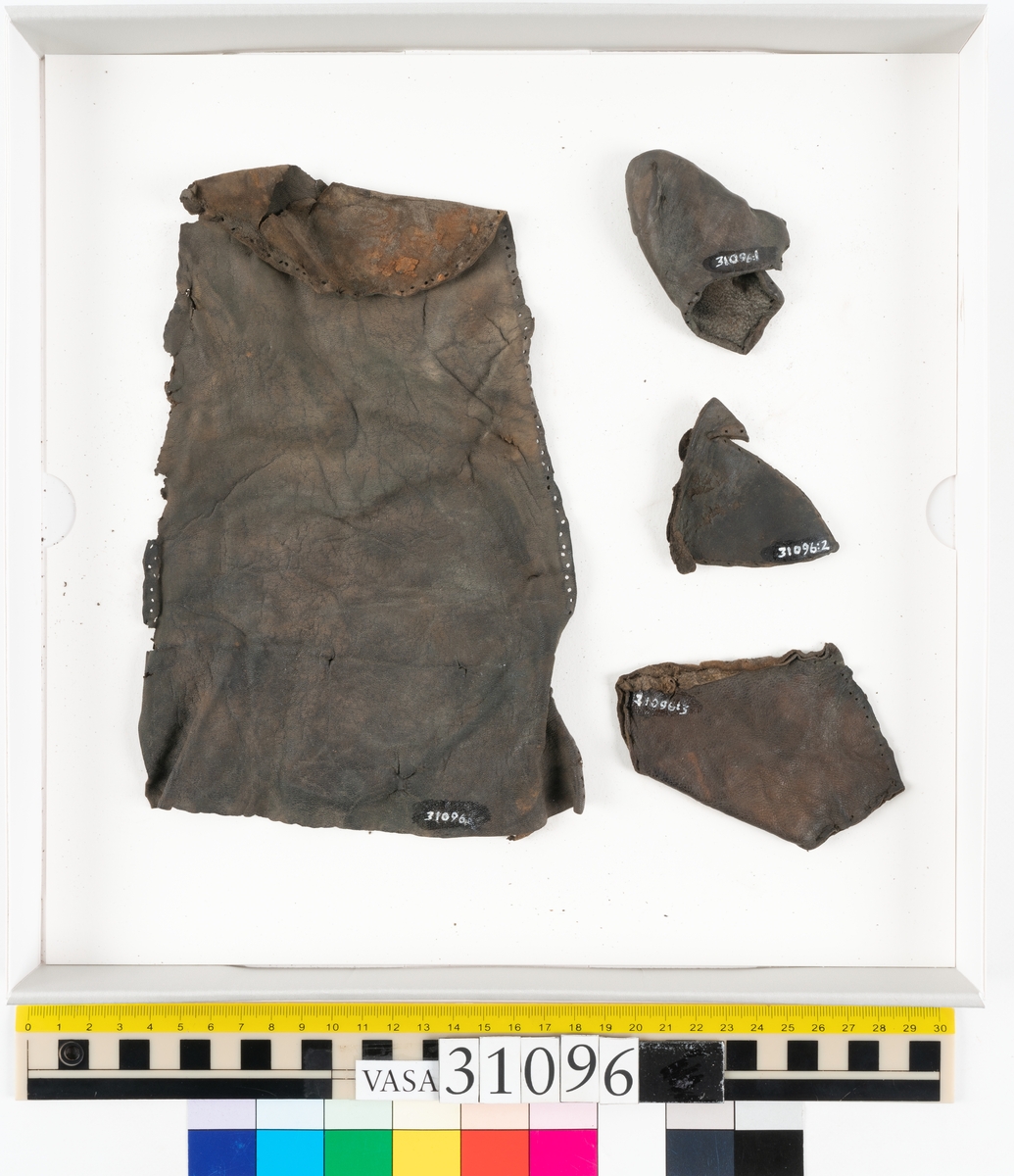 Fyra delar från en tumvante.
Vanten är skuren i två stycken, ett fram- och ett bakstycke, som varit sammanfogat med stygn.
Ett framstycke till en tumvante, fyndnummer 31096. Runt ytterkanterna löper stygnshål. Lädrets kött- och narvsida har släppt från varandra. Vanten har fläckvist rostliknande beläggningar. Lädret är tunt och sprött.
En vanttumme, fyndnummer 31096:1 Längs med ena sidan på vanttummen löper långsmala hål eller öppningar, två och två. Hålen kan härröra från en förstärkning som suttit fästad på vanttummen.
Triangelformat bakstycke till vanttumme, fyndnummer 31096:2. Lädret har skurits ut från vantens bakstycke. Stygnshål är synliga längs med två av sidorna.
Rektangulärt läderstycke, fyndnummer 31096:3. Längs med ytterkanterna löper srygnshål. Lädret är mjukt och narvytan tydlig. Lädret har börjat att spalta sig samt är försett med några rostliknande fläckar. Troligtvis har läderbiten fungerat som en förstärkning och suttit fästad på vantens in - eller utsida.