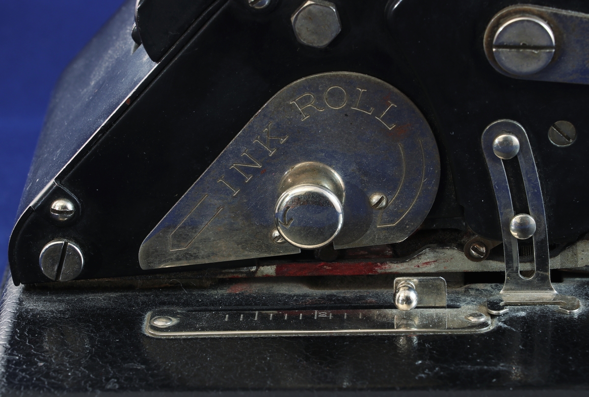 Perforeringsmaskin från 1920-talet. 
Svart platt metallåda med bläcklucka samt lås för öppnings av lådan på undersidan.
Rundad metallblock med åtta reglage för siffror, de tre första reglagen är röda, nästa tre gröna och sista två röda.
Linjal på sidan i metall.
Handtag för tryck.
Platta med modellinfo under rullen.