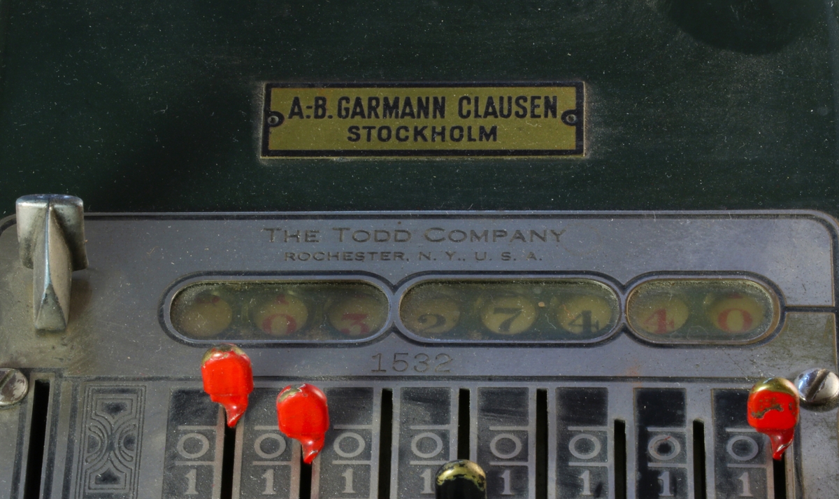 Grön perforeringsmaskin från The Todd Company. Maskinen är fäst på en grön platta. På ovansidan finns åtta sifferrader från 0-9 med reglage, de tre första reglagen är röda, nästa tre svarta och sista två röda. Ett reglage för att återställa sifferreglagen längst till vänster. Knapp i röd bakelit på framsidan och spak med svart bakelithandtag på höger sida.