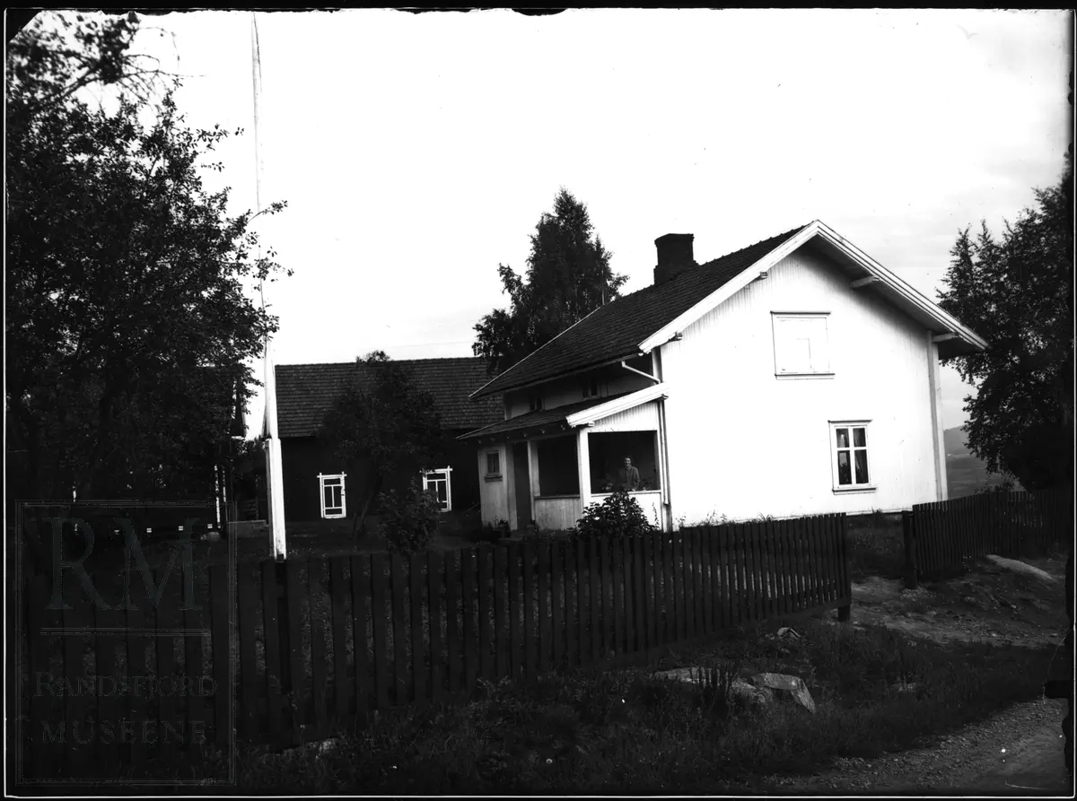 Et bolighus og en liten låve og et stabbur. Stakittgjerde og flaggstang.