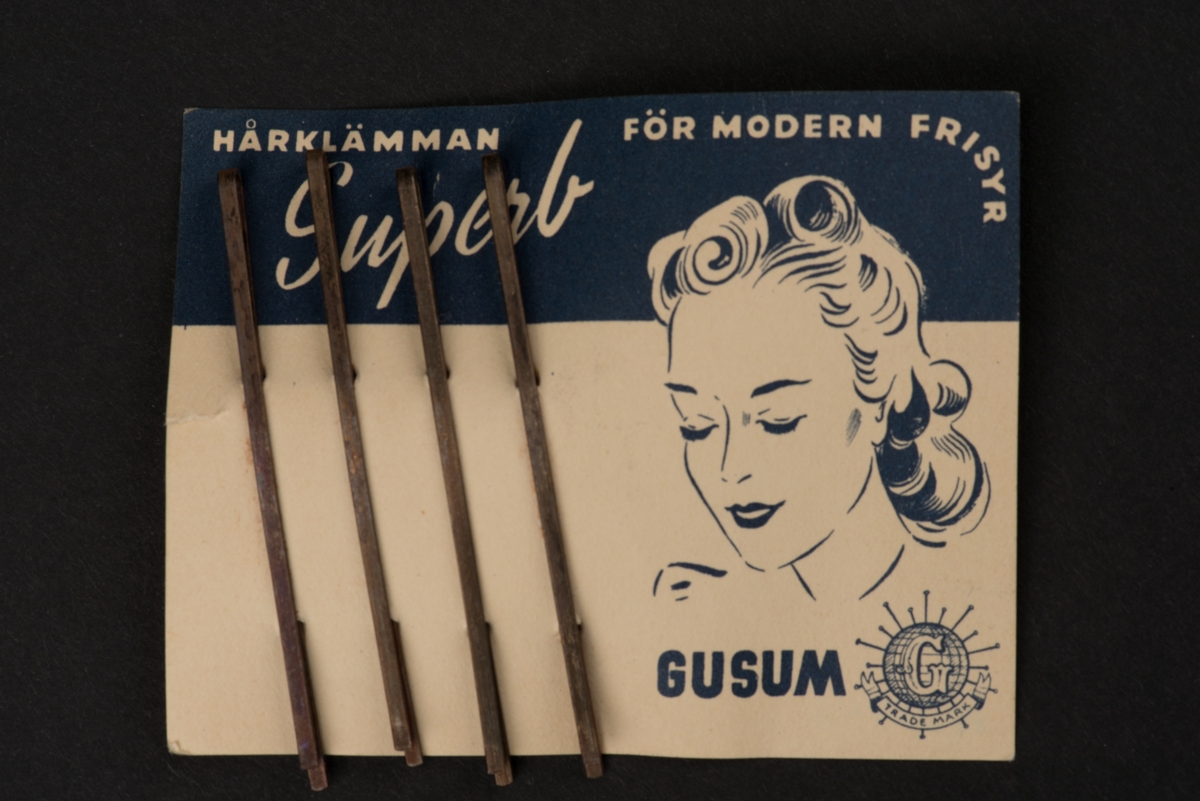 Fyra hårspännen monterade på karta.
Spännena är tillverkade av vikt metallband och lätt böjda. Kartan av papp är i blått och vitt med text och ett avbildat kvinnohuvud med lockig frisyr.