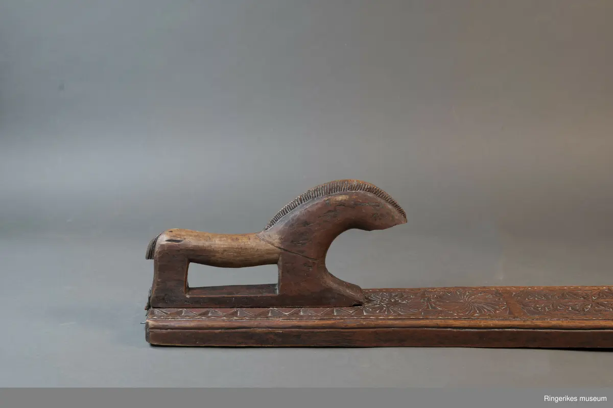 Mørkebrunt mangletre med håndtak forma som en hest. Hesten har man og hale, men mulen er brukket av og mangler. Håndtaket er nedfelt og festa med to nagler fra undersiden. Mangletreet er dekorert med treskurd. Hovedmønsteret består av 5 sirkler hvorav 2 står delvis inni hverandre. 3 av sirklene har seksbladroser inni og 1 har tagger som et sagblad. 1 sirkel har en liten seksbladrose i midten og sagblad tagger rundt. Dekoren består også av to små hjerter og 1814 MD, samt en langsgående sikksakkbord på hver langside og trekantmønster rundt håndtaket.