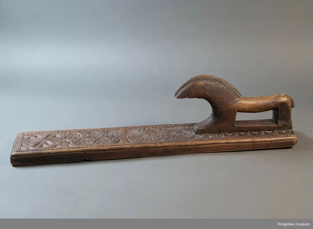 Mørkebrunt mangletre med håndtak forma som en hest. Hesten har man og hale, men mulen er brukket av og mangler. Håndtaket er nedfelt og festa med to nagler fra undersiden. Mangletreet er dekorert med treskurd. Hovedmønsteret består av 5 sirkler hvorav 2 står delvis inni hverandre. 3 av sirklene har seksbladroser inni og 1 har tagger som et sagblad. 1 sirkel har en liten seksbladrose i midten og sagblad tagger rundt. Dekoren består også av to små hjerter og 1814 MD, samt en langsgående sikksakkbord på hver langside og trekantmønster rundt håndtaket.