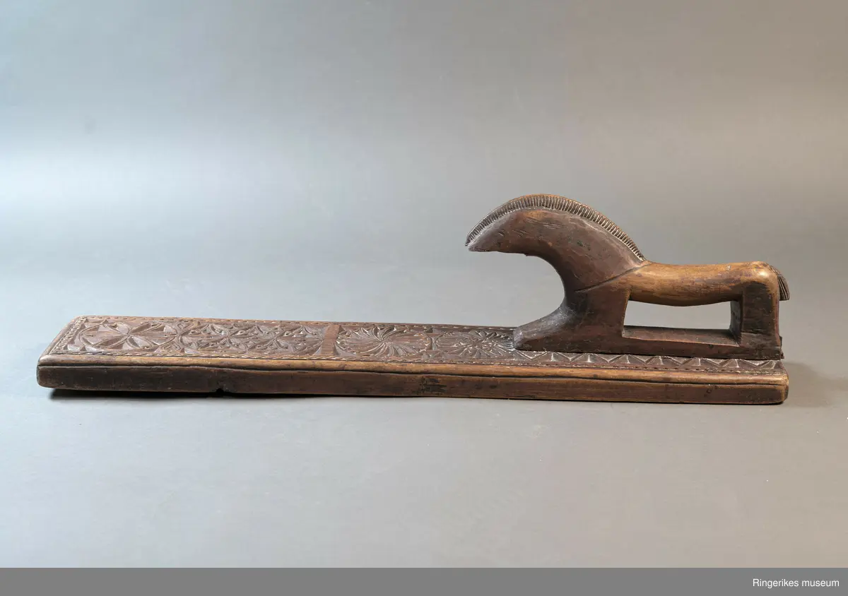 Mørkebrunt mangletre med håndtak forma som en hest. Hesten har man og hale, men mulen er brukket av og mangler. Håndtaket er nedfelt og festa med to nagler fra undersiden. Mangletreet er dekorert med treskurd. Hovedmønsteret består av 5 sirkler hvorav 2 står delvis inni hverandre. 3 av sirklene har seksbladroser inni og 1 har tagger som et sagblad. 1 sirkel har en liten seksbladrose i midten og sagblad tagger rundt. Dekoren består også av to små hjerter og 1814 MD, samt en langsgående sikksakkbord på hver langside og trekantmønster rundt håndtaket.