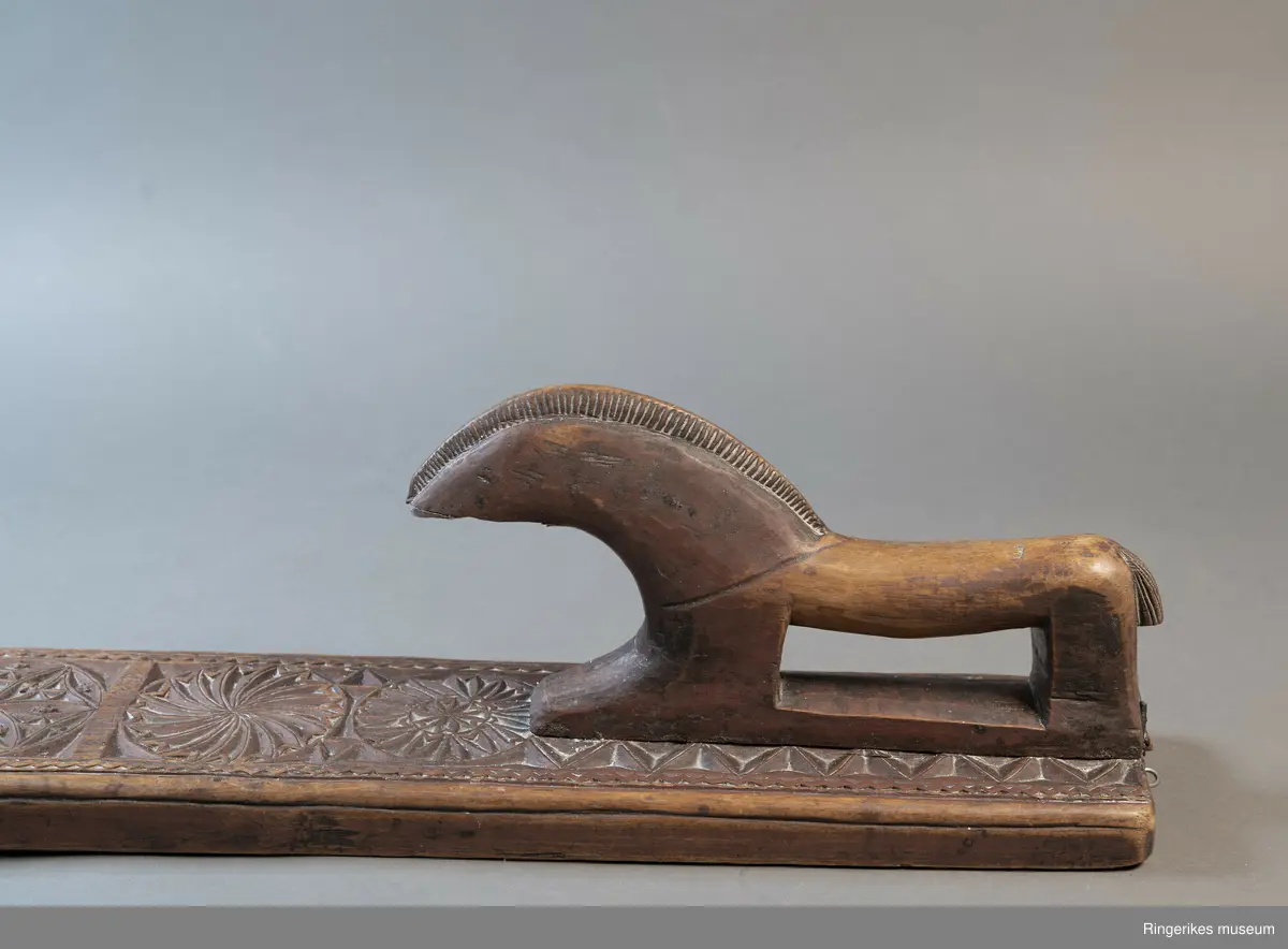 Mørkebrunt mangletre med håndtak forma som en hest. Hesten har man og hale, men mulen er brukket av og mangler. Håndtaket er nedfelt og festa med to nagler fra undersiden. Mangletreet er dekorert med treskurd. Hovedmønsteret består av 5 sirkler hvorav 2 står delvis inni hverandre. 3 av sirklene har seksbladroser inni og 1 har tagger som et sagblad. 1 sirkel har en liten seksbladrose i midten og sagblad tagger rundt. Dekoren består også av to små hjerter og 1814 MD, samt en langsgående sikksakkbord på hver langside og trekantmønster rundt håndtaket.