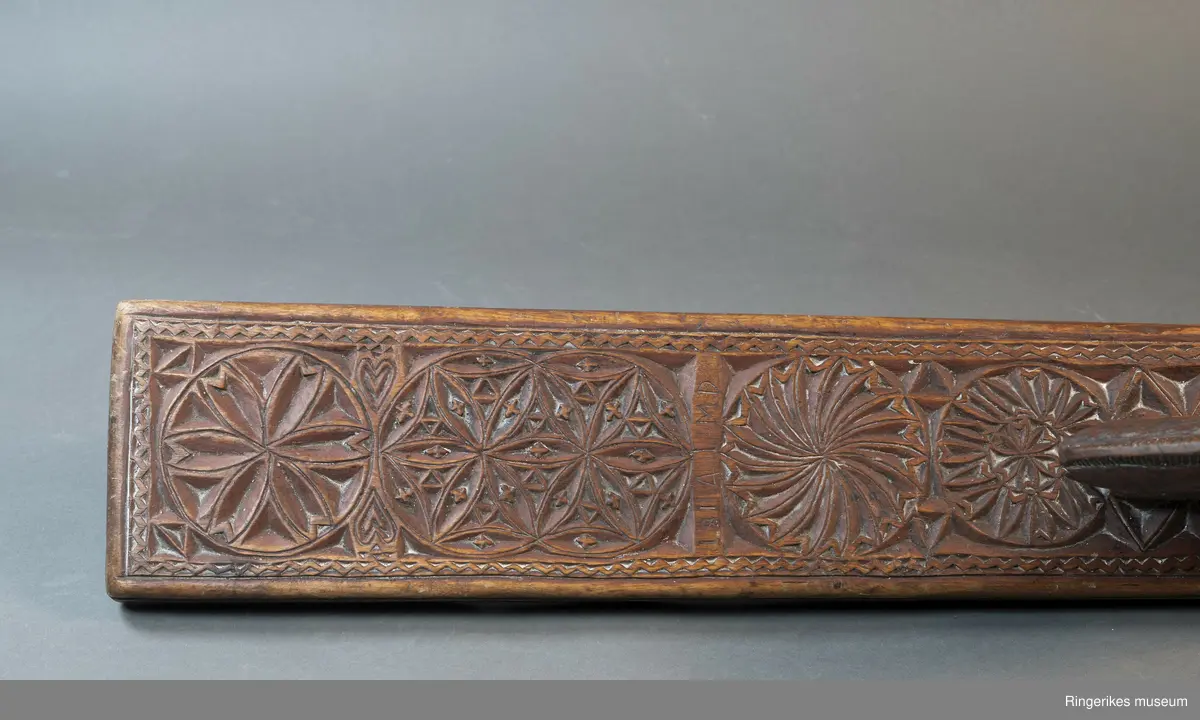 Mørkebrunt mangletre med håndtak forma som en hest. Hesten har man og hale, men mulen er brukket av og mangler. Håndtaket er nedfelt og festa med to nagler fra undersiden. Mangletreet er dekorert med treskurd. Hovedmønsteret består av 5 sirkler hvorav 2 står delvis inni hverandre. 3 av sirklene har seksbladroser inni og 1 har tagger som et sagblad. 1 sirkel har en liten seksbladrose i midten og sagblad tagger rundt. Dekoren består også av to små hjerter og 1814 MD, samt en langsgående sikksakkbord på hver langside og trekantmønster rundt håndtaket.