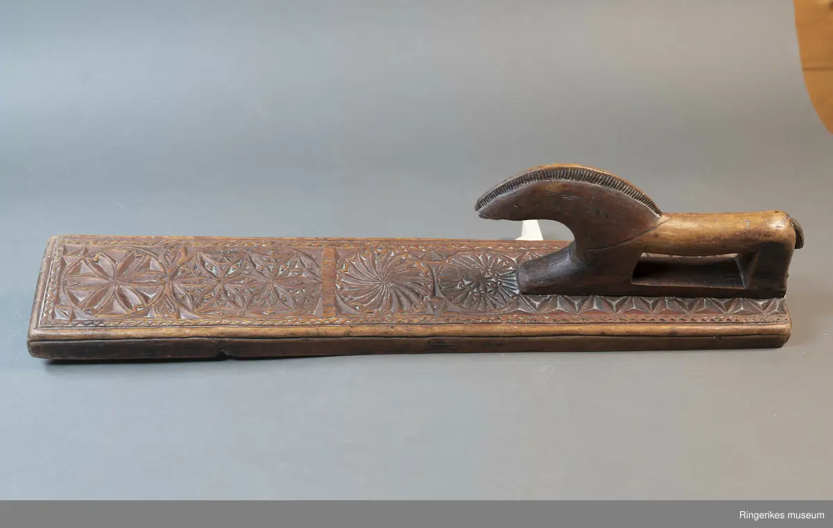 Mørkebrunt mangletre med håndtak forma som en hest. Hesten har man og hale, men mulen er brukket av og mangler. Håndtaket er nedfelt og festa med to nagler fra undersiden. Mangletreet er dekorert med treskurd. Hovedmønsteret består av 5 sirkler hvorav 2 står delvis inni hverandre. 3 av sirklene har seksbladroser inni og 1 har tagger som et sagblad. 1 sirkel har en liten seksbladrose i midten og sagblad tagger rundt. Dekoren består også av to små hjerter og 1814 MD, samt en langsgående sikksakkbord på hver langside og trekantmønster rundt håndtaket.