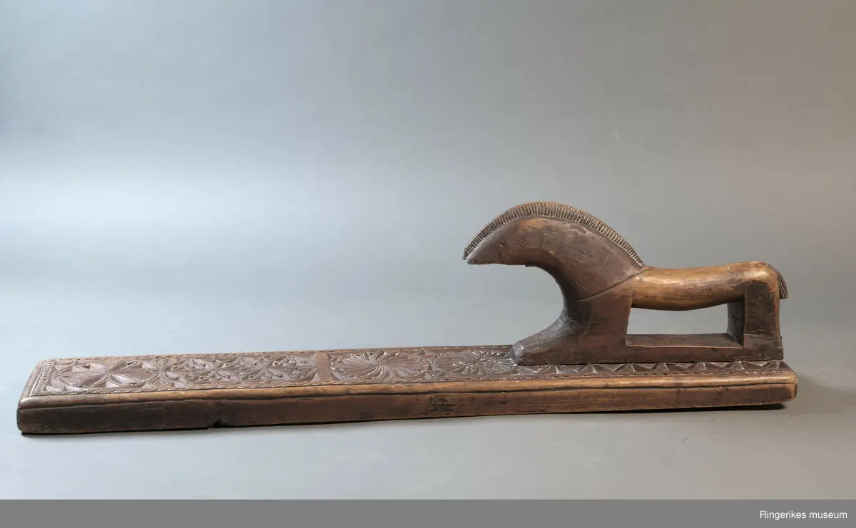 Mørkebrunt mangletre med håndtak forma som en hest. Hesten har man og hale, men mulen er brukket av og mangler. Håndtaket er nedfelt og festa med to nagler fra undersiden. Mangletreet er dekorert med treskurd. Hovedmønsteret består av 5 sirkler hvorav 2 står delvis inni hverandre. 3 av sirklene har seksbladroser inni og 1 har tagger som et sagblad. 1 sirkel har en liten seksbladrose i midten og sagblad tagger rundt. Dekoren består også av to små hjerter og 1814 MD, samt en langsgående sikksakkbord på hver langside og trekantmønster rundt håndtaket.