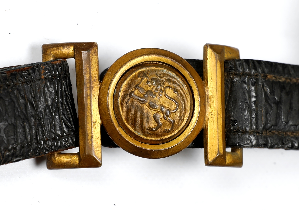 Norsk bandolær produsert mellom 1844 og 1905. Svartfarget lær med påsydd ullstoff på undersiden. Spenne i messing med løvefigur til å feste beltet foran. Bandolæret er montert feil vei og det kan mangle en kartusjveske.