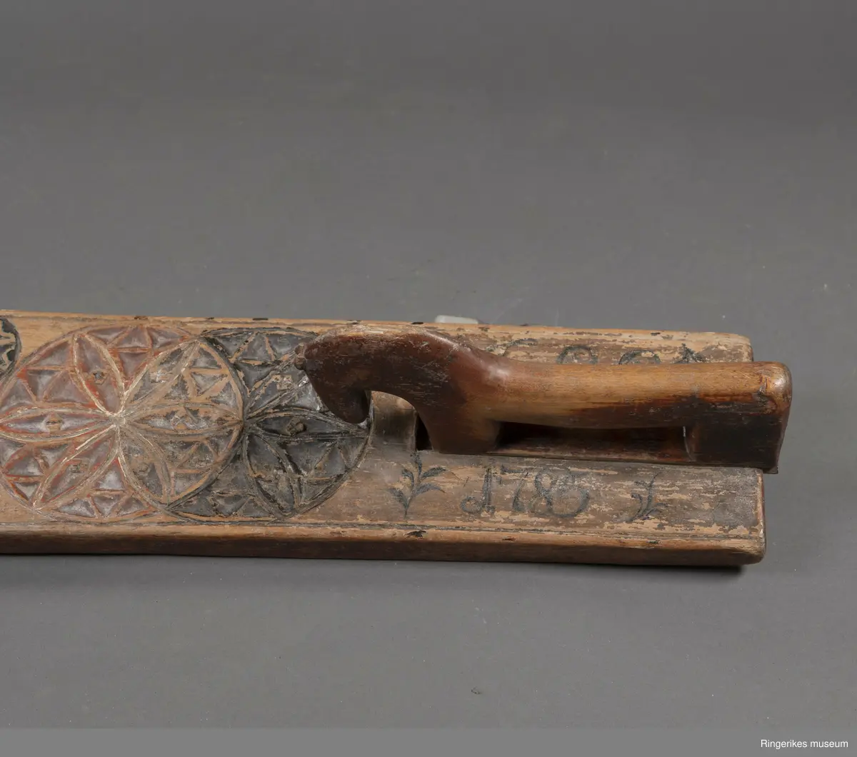 Mangletre med innfelt håndtak formet som en hest. Mangletreet er dekorert med treskurd hvor hovedmønsteret består av 4 sirkler med seksbladroser inni. 2 av sirklene står delvis inni hverandre. 4 små sirkler med åttebladroser er plassert mellom de store sirklene. Sirklene har opprinnelig vært malt og fremstår i dag som røde og svarte. På den ene siden ved håndtaket er det påmalt 1785 og på den andre siden GGD.