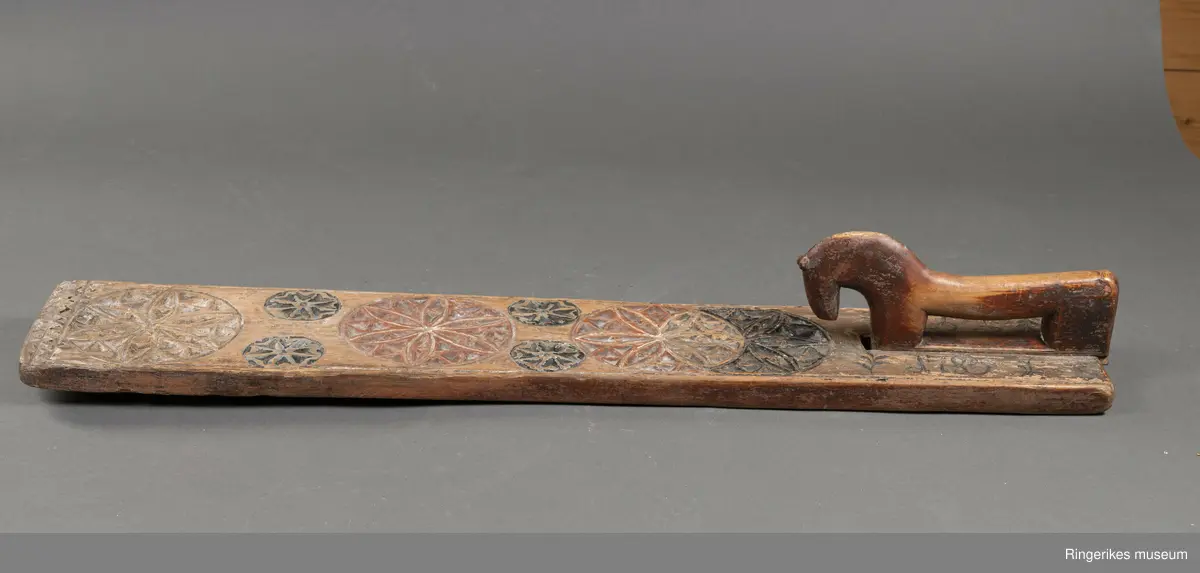 Mangletre med innfelt håndtak formet som en hest. Mangletreet er dekorert med treskurd hvor hovedmønsteret består av 4 sirkler med seksbladroser inni. 2 av sirklene står delvis inni hverandre. 4 små sirkler med åttebladroser er plassert mellom de store sirklene. Sirklene har opprinnelig vært malt og fremstår i dag som røde og svarte. På den ene siden ved håndtaket er det påmalt 1785 og på den andre siden GGD.