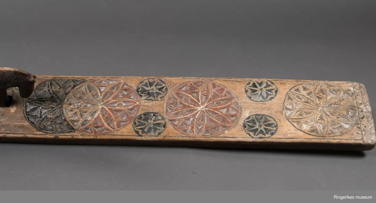 Mangletre med innfelt håndtak formet som en hest. Mangletreet er dekorert med treskurd hvor hovedmønsteret består av 4 sirkler med seksbladroser inni. 2 av sirklene står delvis inni hverandre. 4 små sirkler med åttebladroser er plassert mellom de store sirklene. Sirklene har opprinnelig vært malt og fremstår i dag som røde og svarte. På den ene siden ved håndtaket er det påmalt 1785 og på den andre siden GGD.