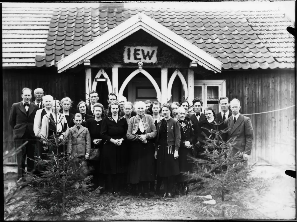 Begravelse. En gruppe står utenfor et hus. Over inngangen står bokstavene IEW.