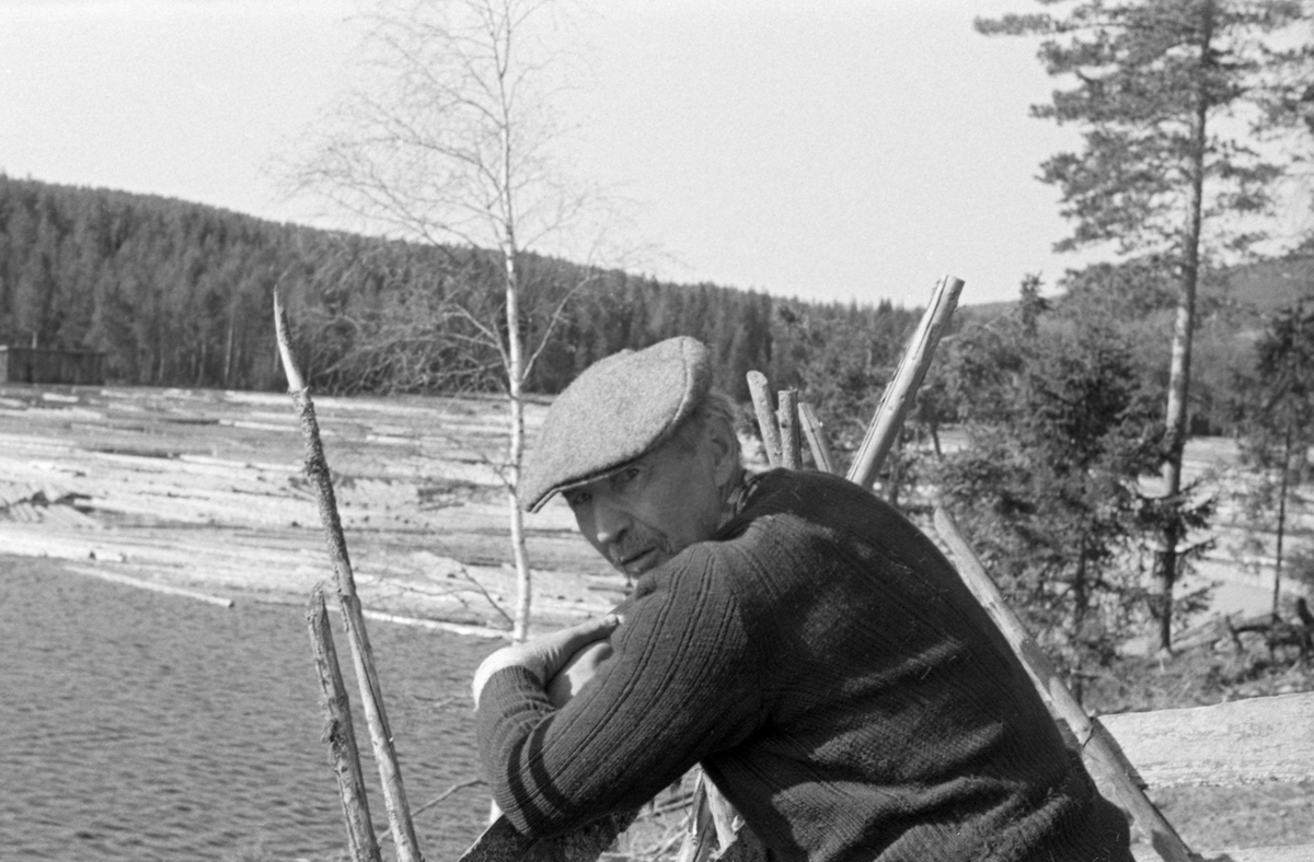 Portrett av tømmerfløteren John Bergsødegården (1887-1957) fra Mo i Nord-Odal i Hedmark.  Bergsødegården er fotografert ved en skigard eller et vedreis, iført en tynn, mørk genser og med ei grålig sixpencelue på hodet.  I bakgrunnen ser vi et vannspeil, antakelig Storsjøen i Odalen, med en del barket løstømmer i vannskorpa.