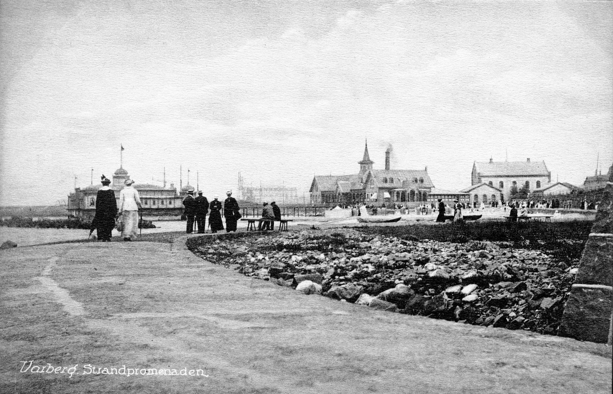 Vykort, "Varberg. Strandpromenaden". Strandpromenaden vid Barnens badstrand med varm- och kallbadhusen samt tullhuset. Personer promenerar mot badhusen, hamnen.