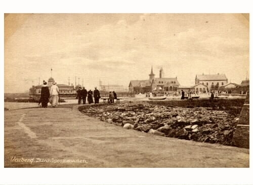 Vykort, "Varberg. Strandpromenaden". Strandpromenaden vid Barnens badstrand med varm- och kallbadhusen samt tullhuset. Personer promenerar mot badhusen, hamnen.
