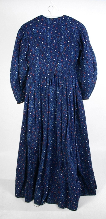 Klänning i blå bottenfärg, vattrat mönster, prickar i gult, rött och grönt. Slätt figurskuret liv med insydda fjädrar. Valben i ryggsöm och insnitt fram. Knäppning framtill med 13 st hyskor och hakar, varav flera saknas. Ingen krage. Isydd ärm med lagda veck över axeln. Passpoal i sömmen. Smalt markerat ryggstycke med svarta passpoalmarkerade sömmar. En vid ärm som smalnar vid handleden, vilken är kantad med svarta bomullsband, vilka nu är gröna. Kjolen har lagda veck runt om, sprund mitt fram. Sydd av 4 hela våder och en kilskuren våd. Sparkband av samma bruna linnelärft som livets foder. Vänster ärm är fodrad med brunt kattuntyg med mönster av vita prickar på brun botten, däröver brandgula blommor och streckliknande mönster. Klänningen är lagad på flera ställen och hålig.