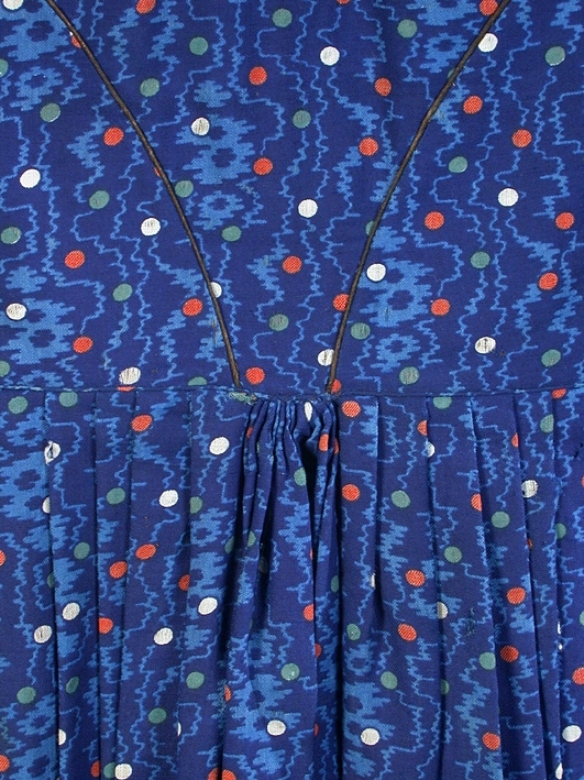 Klänning i blå bottenfärg, vattrat mönster, prickar i gult, rött och grönt. Slätt figurskuret liv med insydda fjädrar. Valben i ryggsöm och insnitt fram. Knäppning framtill med 13 st hyskor och hakar, varav flera saknas. Ingen krage. Isydd ärm med lagda veck över axeln. Passpoal i sömmen. Smalt markerat ryggstycke med svarta passpoalmarkerade sömmar. En vid ärm som smalnar vid handleden, vilken är kantad med svarta bomullsband, vilka nu är gröna. Kjolen har lagda veck runt om, sprund mitt fram. Sydd av 4 hela våder och en kilskuren våd. Sparkband av samma bruna linnelärft som livets foder. Vänster ärm är fodrad med brunt kattuntyg med mönster av vita prickar på brun botten, däröver brandgula blommor och streckliknande mönster. Klänningen är lagad på flera ställen och hålig.