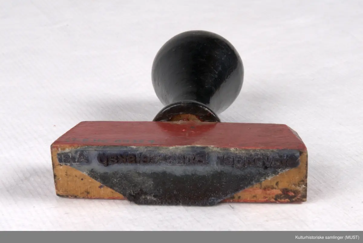 Stempel med rektangulært stempelhode av rødmalt tre med metallstempel festet under, og med sortmalt håndtak av dreid tre. Stempelet er delvis utydelig. Det er plass til å fylle inn en signatur mellom linjene i stempelet.