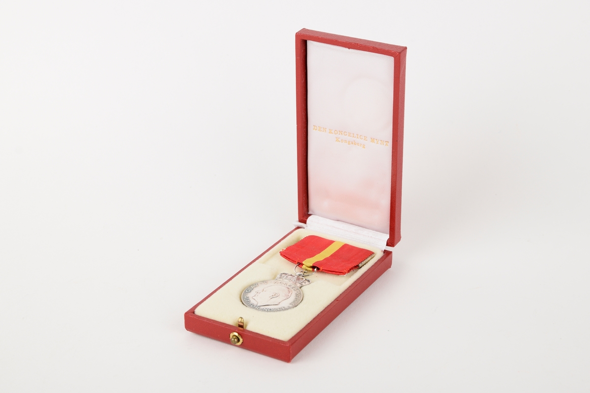 Et eksemplar av "Kongens fortjenstmedalje" i originalt etui. 

Sirkulær medalje med ordensbånd, oppbevart i rektangulært etui. Etuiet har motiv av en krone i sølv på lokket.
