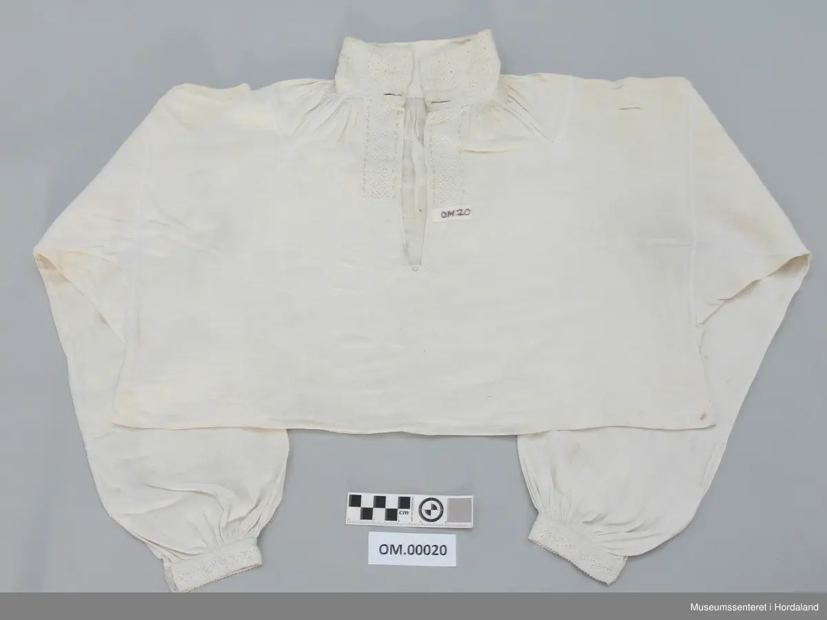 Kvit linskjorte med rynking rundt hals og armar. Krage, armmansjettar og stolpe er sydd med kvit lintråd i enkel uttrekkssaum. Kilar/stoffstykke under armane og på skuldrane. Skjorta er ganske kort i livet. Ikkje splittar i sidene.