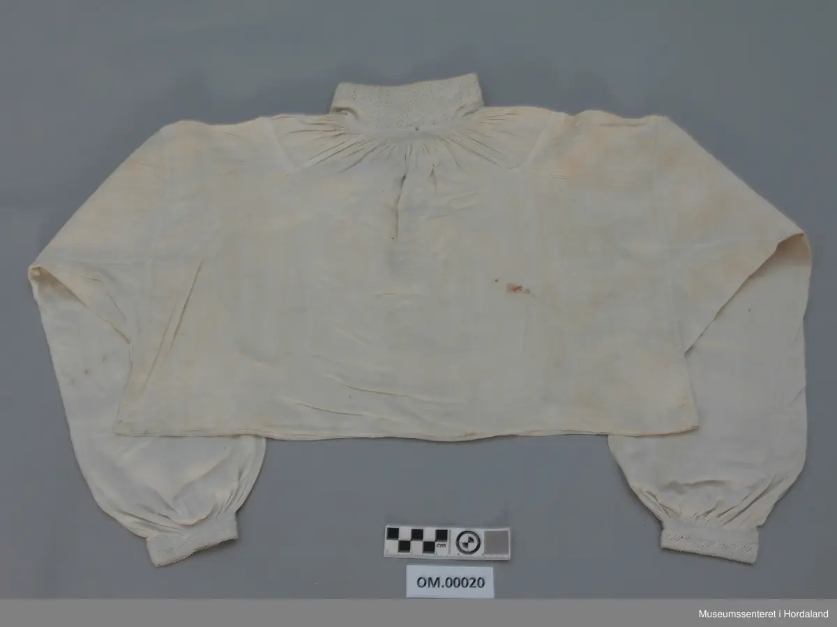 Kvit linskjorte med rynking rundt hals og armar. Krage, armmansjettar og stolpe er sydd med kvit lintråd i enkel uttrekkssaum. Kilar/stoffstykke under armane og på skuldrane. Skjorta er ganske kort i livet. Ikkje splittar i sidene.