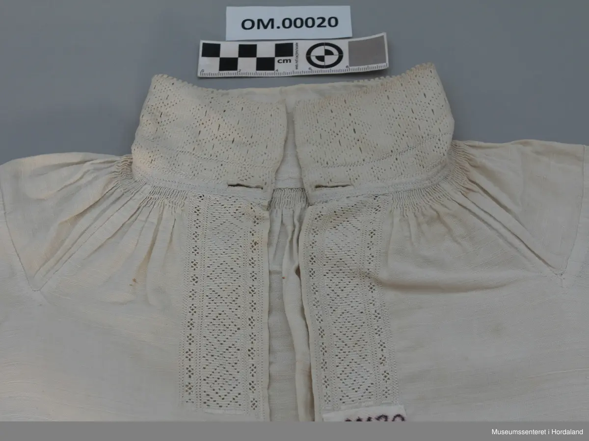 Kvit linskjorte med rynking rundt hals og armar. Krage, armmansjettar og stolpe er sydd med kvit lintråd i enkel uttrekkssaum. Kilar/stoffstykke under armane og på skuldrane. Skjorta er ganske kort i livet. Ikkje splittar i sidene.