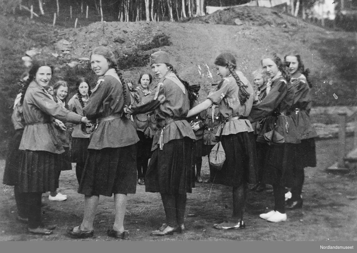 speiderpiker. fra leir ved Trondenes. en gruppe pikespeidere holder hverandre i hendenenen og danner en ring. i bakgrunnen jordvoll. Helga og Ole Vatten er besteforeldre til giver av bildet, og Helga Vatten var formann i byggekomiteen for byggingen av ny speiderhytte etter andre verdenskrig.