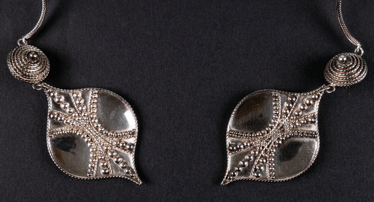 Ashwa, smycke i silver av Rosa Taikon.
Öronsmycke i form av två silverlöv med granalieornamentik, vilka via varsin granelerad kula hänger i en dubbel bygel att sätta över huvudet.