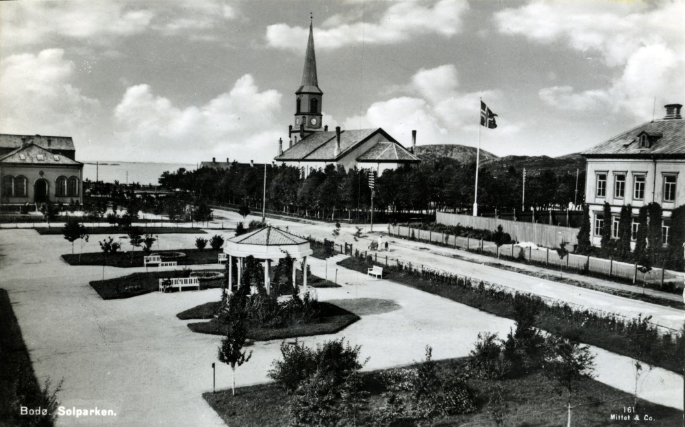 Postkort med motivi fra Bodø. Solparken i forkant og museet og Bodø kirke i bakgrunnen.