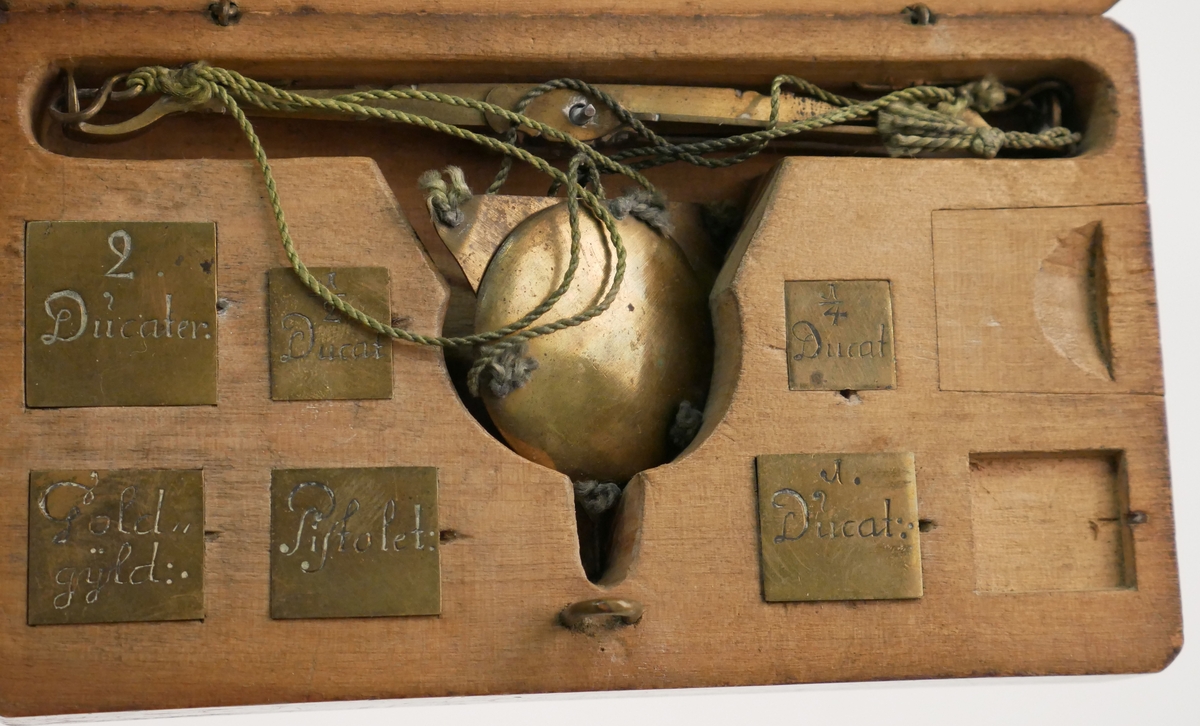 Träask med våg samt vikter. Vikterna märkta: 2 Ducater, 1 Ducat, 1/2 Ducat, 1/4 Ducat, Pistolet och Gold Gÿld. På askens lock står "B.W. Meijer Anno 1733". En vikt saknas.