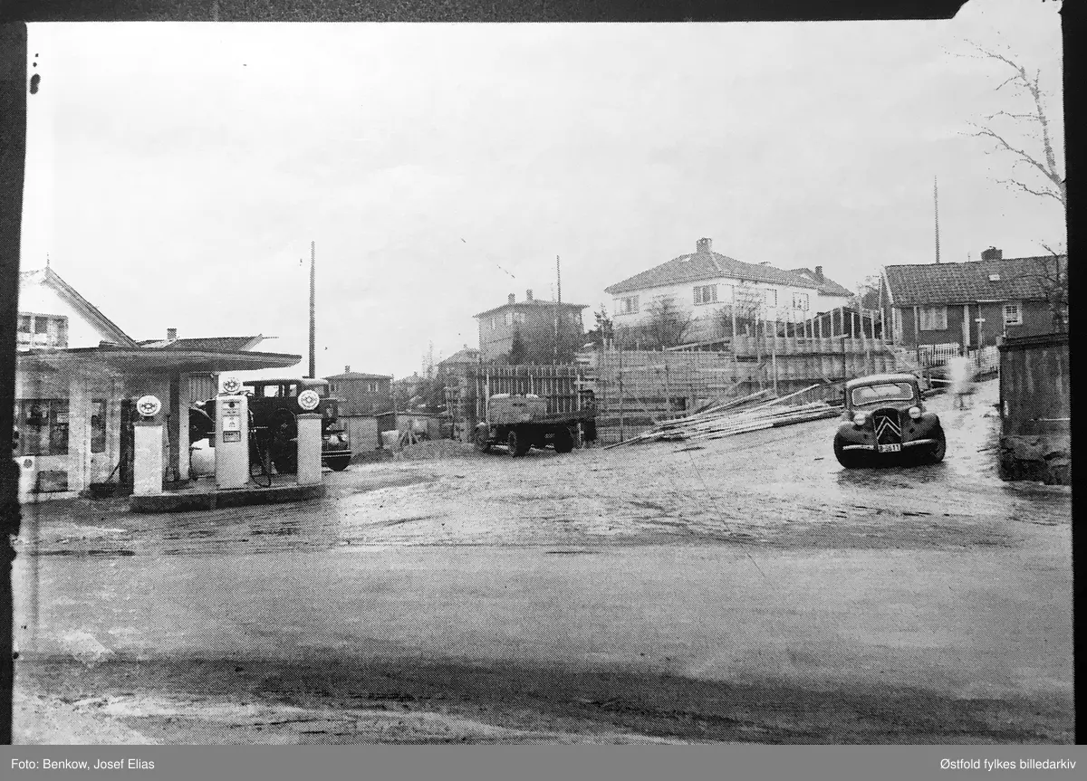Bensinstasjon i Klostergata 12 i Moss, selger av bensinmerket NOR (Østlandet). Oslo Sporveier sto bak, men NOR ble fusjonert inn i Nor & Vacuum ca. 1952, senere ble det Mobil. Biler med skilt: Lastebil B-8610, personbil B-6611.
Bilen til høyre er en klassisk Citroën Traction Avant, som kom ny i 1935 og var i produksjon til 1955, 
Bestiller av bildet  er ingeniør Stenmark.
Martebakken til høyre. Gubberogata til venstre. Gubberogata 1 under oppføring.