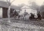 Nött men unikt fotografi från Tullgarn omkring förra sekelskiftet. Bilen, en Peugeot modell 28, inköptes år 1900 av kronprins Gustaf för sina resor mellan Stockholm och kungsgården Tullgarn som han vid tiden disponerade. Bilens förare har av John Nerén (förf. Automobilens historia) antagits vara verkmästare G Behmer alternativt Brehmer men har för denna uppgift inte närmare identifierats. Kusken får antas vara Klas Robert Thorell som denna tid var anställd vid Tullgarn. Byggnaden i bakgrunden är Tullgarns värdshus.