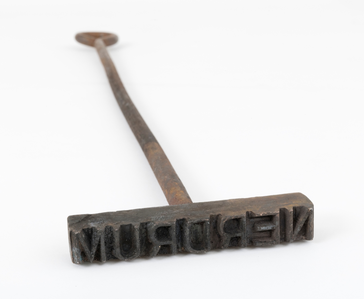 Svijernstempel, brukt til å merke objekter av tre. Sjølve stempelet er lagd av et rektangulært jernstykke (9,6 X 1,6 centimeter) med trapesformet tverrsnitt, Langs den smaleste ytterkanten på det rektangulære jernstykket er det frest inn bokstaver som når man ser stempelet fra undersida leses som «MURDREN» - en speilvendt versjon av navnet «NERDUM». Her refereres det til Nerdrum bruk, et sagbruk i Fet kommune på Nedre Romerike. Vinkelrett på midten av oversida av det rektangulære jernstykket er det innskrudd ei drøy 0,9 centimeter tjukk jernstang, som er rett i 44 centimeters lengde, men som i overenden er bøyd til en opphengsring.

Dette svijernstempelet ble avlevert til Norsk Skogbruksmuseum i ei trekasse med mange slike stempler. Navnene på mange av stemplene refererer til tømmeroppkjøpere og skogbaserte industribedrifter. Gjenstanden er sannsynligvis ett av mange svijernstempler som ble brukt på skaftene på merkeøksene som ble brukt ved innmåling av fløtingstømmer.


Det er 53 svijernstempler med registreringsnumre fra og med SJF.08403 til og med SJF.08456. Alle ligger samlet i ei trekasse.