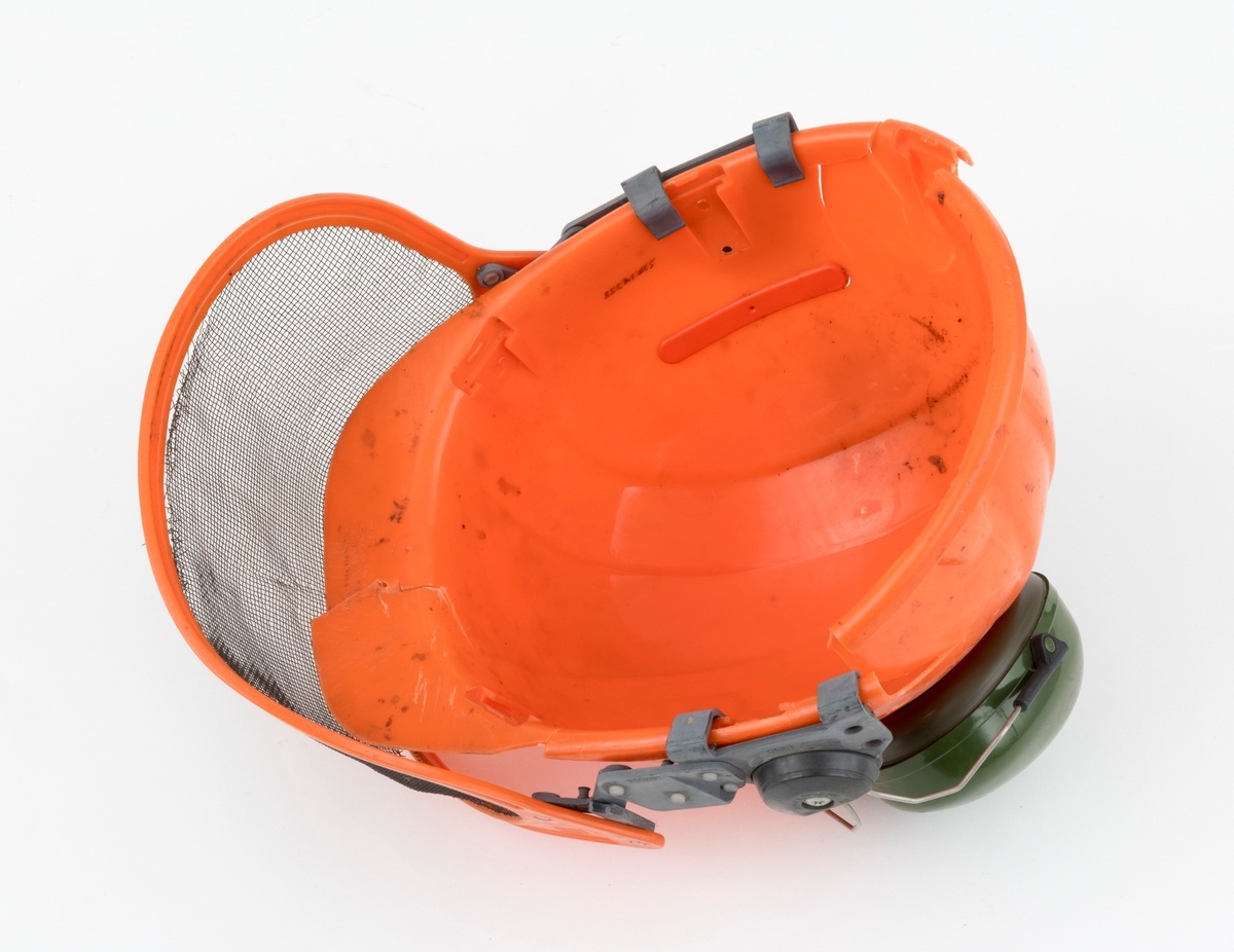 Hjelm, vernehjelm, hoggerhjelm, skogshjelm, for bruk under skogsarbeid. Hjelmen er støpt i hardplast. Den er utsyrt med visir og hørselvern (øreklokker). Det er flere sprekkdannelser hjelmens plastskall. Visirets netting har røket på to steder ved innfestingen i plastramma. Regulerbare reimer for tilpasning til brukerens hode mangler. 
Visir og hørselvern (øreklokker) er festet i samme "plastbrakett". Hørselvernene (øreklokkene) er regulerbare og kan svinges opp på hjelmen med en enkel håndbevegelse. (Hørselvern og visir kan for registrator se ut til å være av noe nyere dato enn selve hjelmen, men det er ikke bekreftet. ) Hørselvernene har for øvrig skomgummpolstring som ligger an mot brukerens ører. 
Hjelmen har to luftehull på hver side som kan åpnes og lukkes med vridbare plastklaffer. Klaffene betjenes fra innsiden.