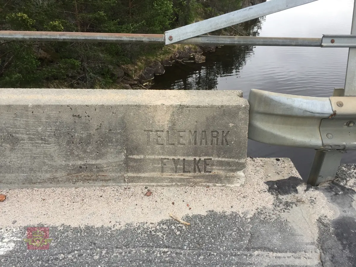 Bruskilt i betong støpt inn i autovernet på Nobbesund bro fv. 276 hovedparsell 1 km 4,8. Fellevegen med teksten: Aust-Agder fylke. Registrert for Statens vegvesen av Næs Jernverksmuseum i 2012 som del av prosjekt med registrering av milepæler og grensemerker mm. 
Se flere bilder i Relasjoner.