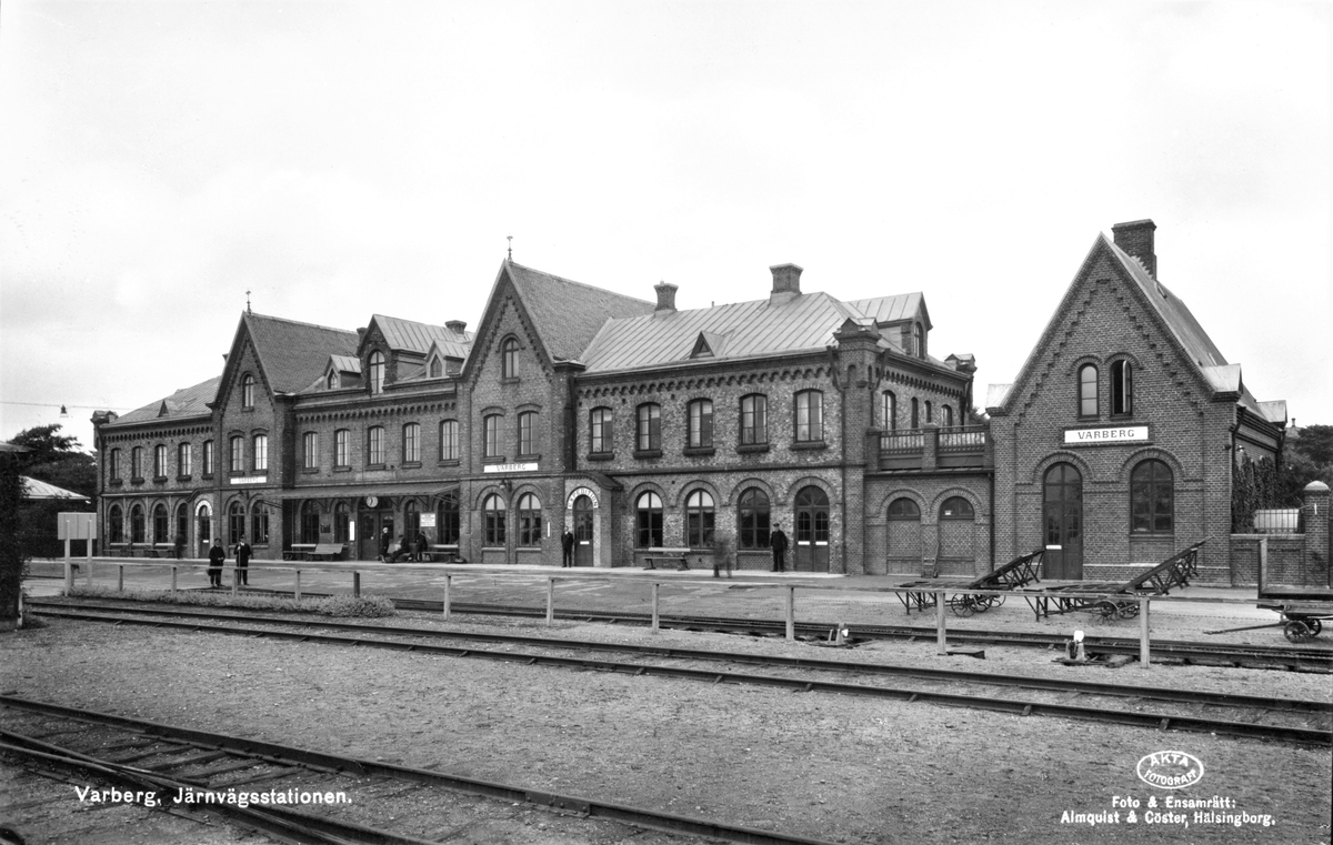 Vykort, "Varberg. Järnvägsstationen." märkt med "Äkta fotografi". Bilden visar stationsbyggnaden sedd från spårsidan. Klockan visar på strax efter halv tolv, både resenärer och järnvägspersonal syns vid byggnaden, till höger väntar ett antal tomma kärror på gods. Utanför expeditionens entré står kanske föreståndaren själv. Här ser man tydligt stationsbyggnadens olika tillbyggnader: i mitten står den urpsrungliga huskroppen med sina två höga sadeltak; på ömse sidor tillkom symmetriska tillbyggnader 1893; längst till höger (i söder) uppfördes 1923 en godsexpedition med matchande högt sadeltak och fasadkrenelering, men i avvikande rött tegel istället för gult.