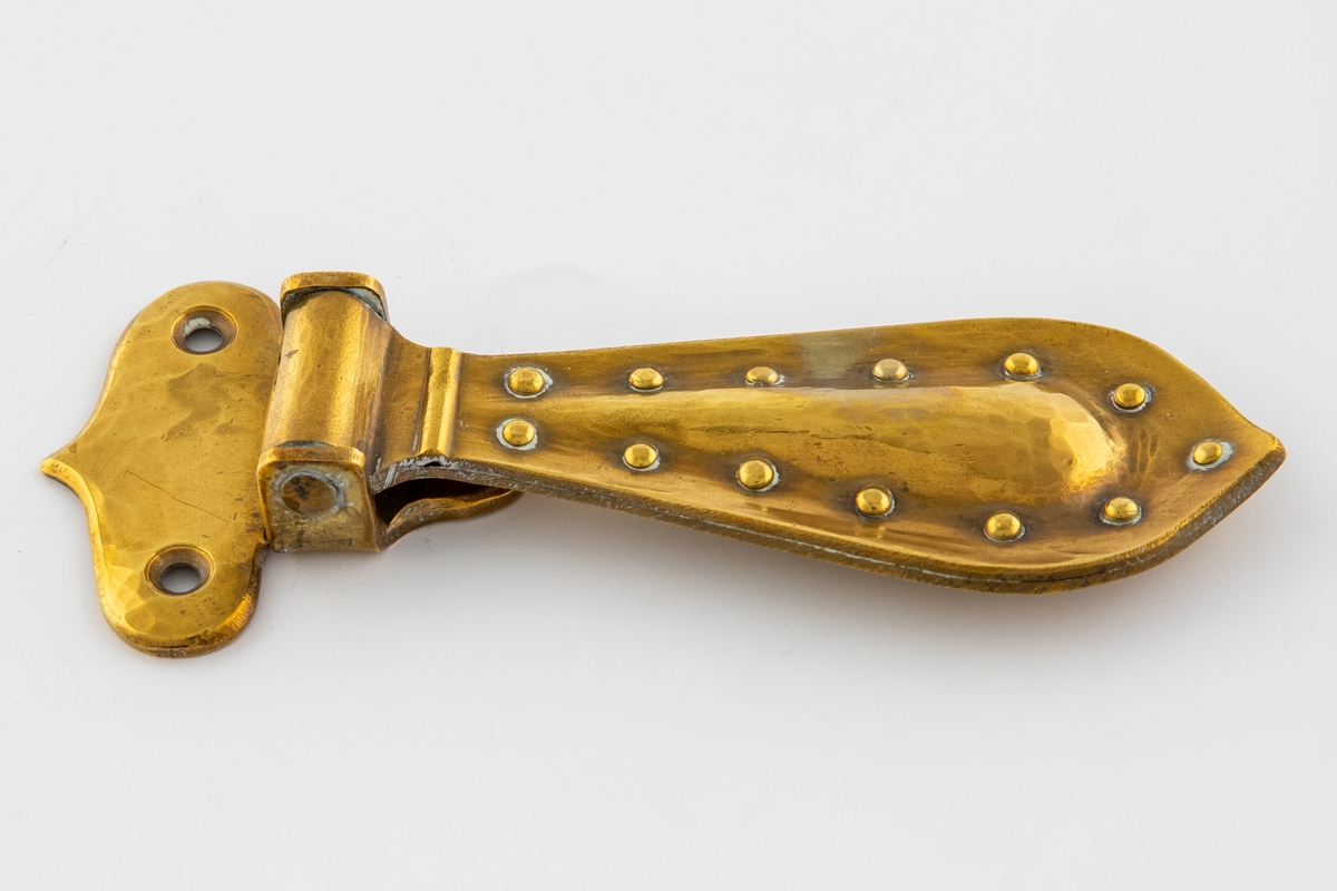 Hamret beslag i messing med hengende håndtak. Beslaget er lite og har en avrundet T-form med tre festehull. Håndtaket er større og dråpeformet, det er festet i en tverrstang og henger nedover. Håndtaket består av to deler som buler utover, de er hamret sammen og festet med messingnitter langs kanten.