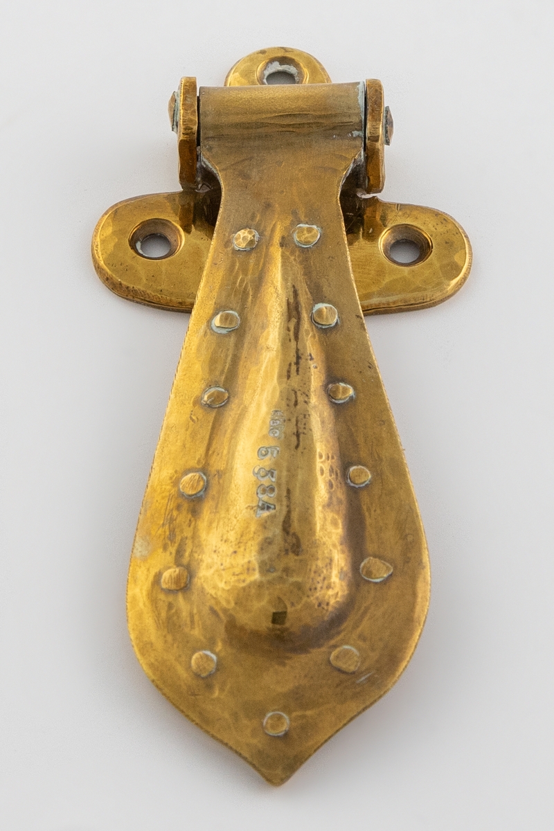 Hamret beslag i messing med hengende håndtak. Beslaget er lite og har en avrundet T-form med tre festehull. Håndtaket er større og dråpeformet, det er festet i en tverrstang og henger nedover. Håndtaket består av to deler som buler utover, de er hamret sammen og festet med messingnitter langs kanten.