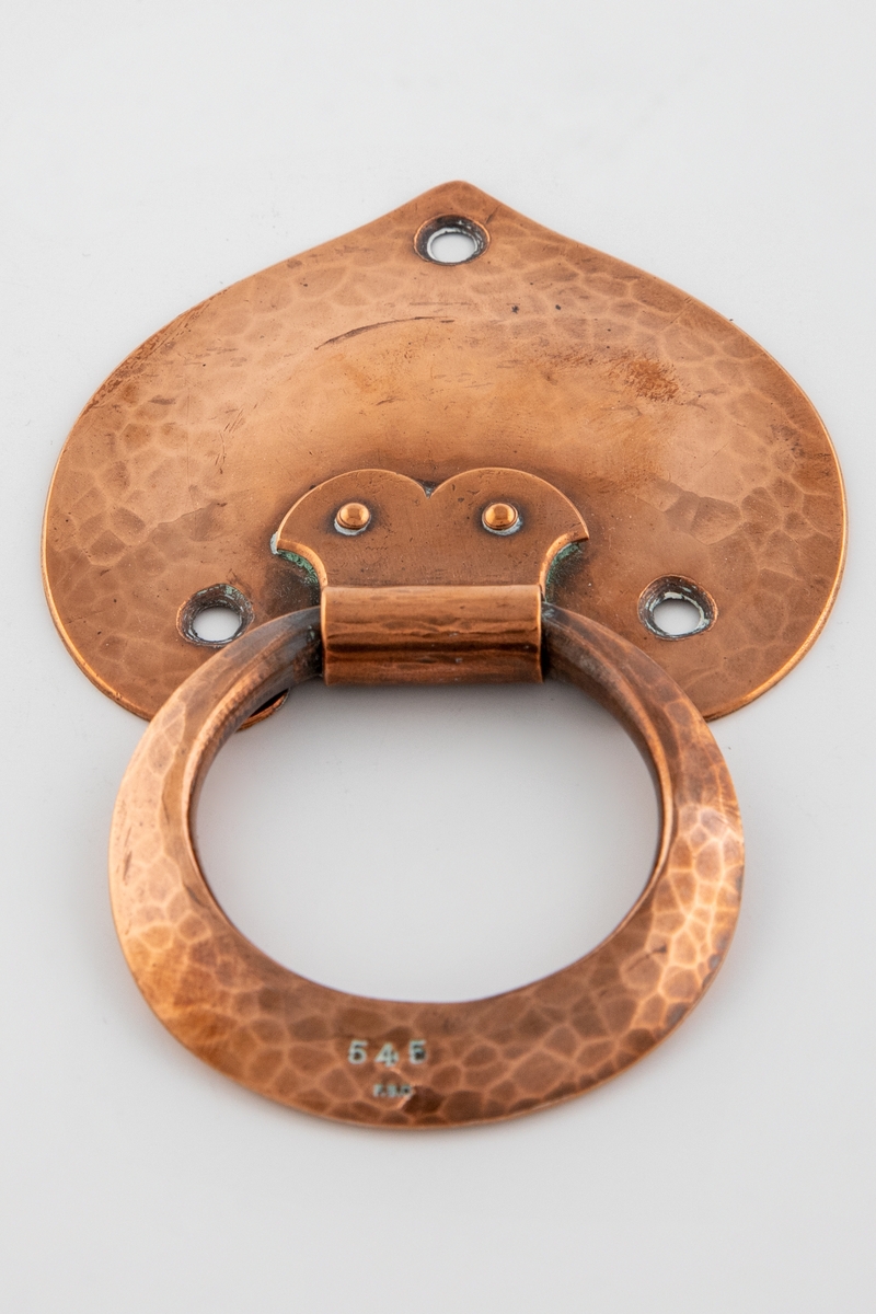 Hamret beslag i kobber. Selve beslaget er hjerteformet med tre festehull og en svak heving i midten. Håndtaket er ringformet, der den nedre delen er svakt utflatet. Ringen er festet på toppen av beslaget i et rørformet feste med to fliker, som igjen er festet med to nitter.