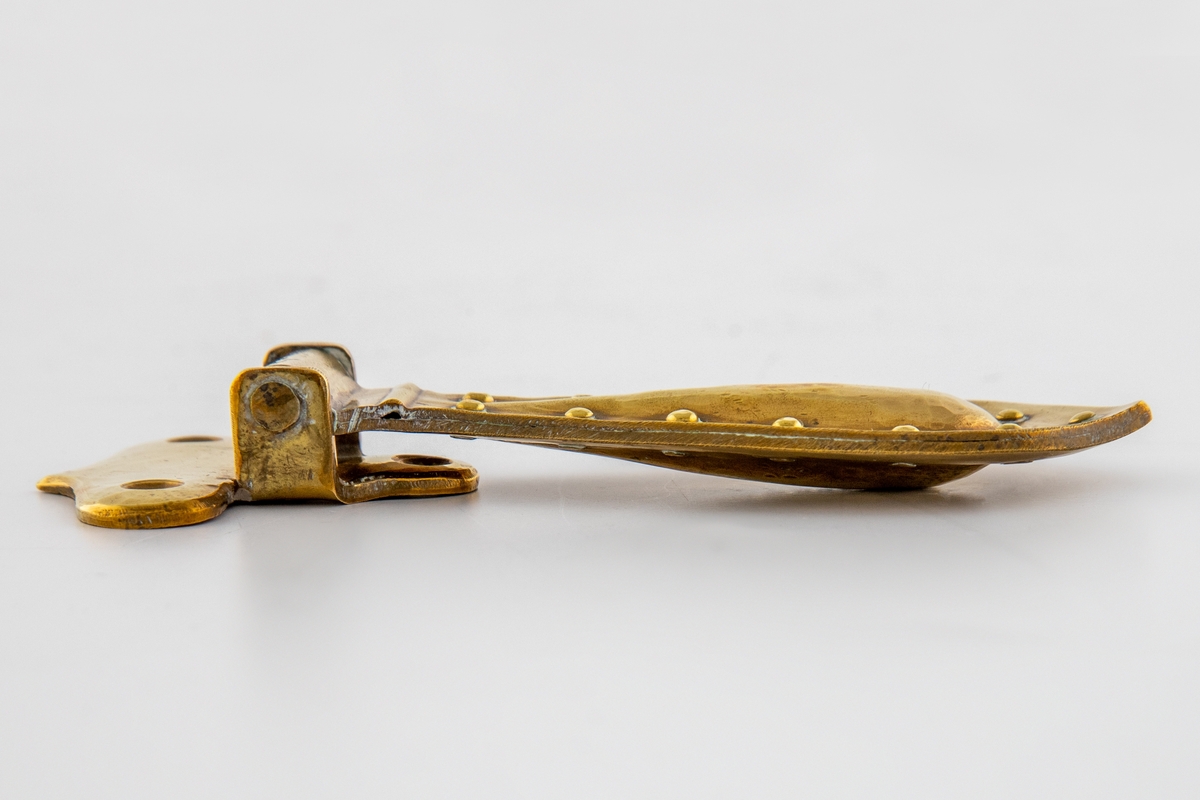 Hamret beslag i messing med hengende håndtak. Beslaget er lite og har en avrundet T-form med tre festehull. Håndtaket er større og dråpeformet, det er festet i en tverrstang og henger nedover. Håndtaket består av to deler som buler utover, de er hamret sammen og festet med messingnitter langs kanten.