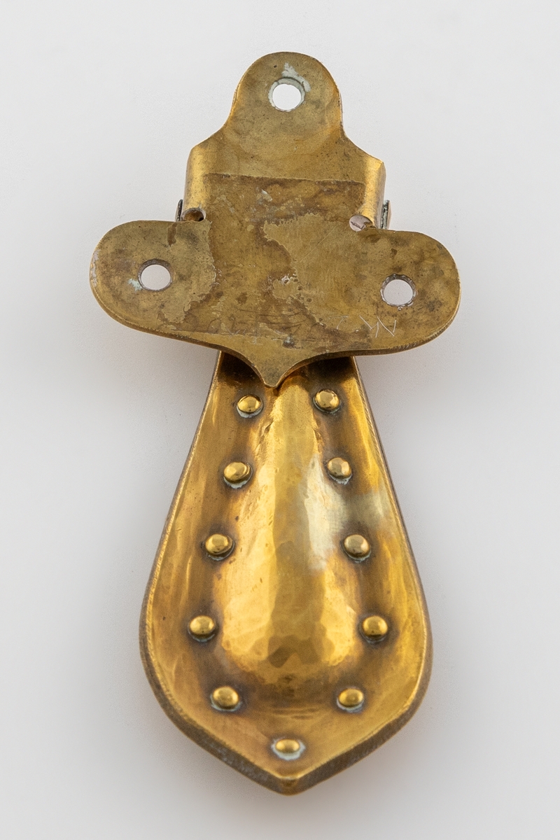 Hamret beslag i messing med hengende håndtak. Beslaget er lite og har en avrundet T-form med tre festehull. Håndtaket er større og dråpeformet, det er festet i en tverrstang og henger nedover. Håndtaket består av to deler som buler utover, de er hamret sammen og festet med messingnitter langs kanten.