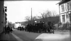 Ett liktåg med vagn dragen av hästar på Brahegatan på väg söderåt. Man passerar den avlidnes hem som skymtar till höger.