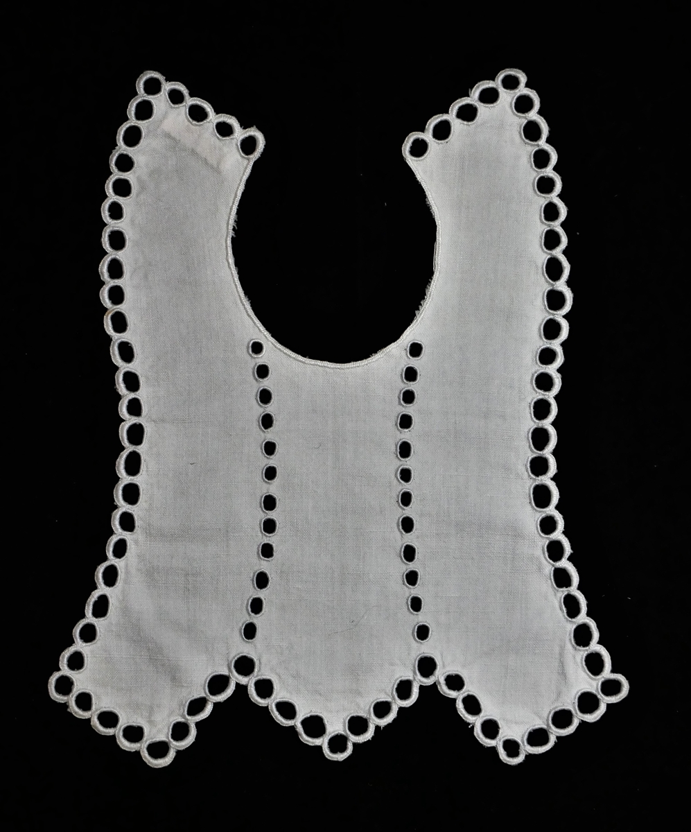 Spebarnssmekke forma som krage bak. Framme utoversvinga sider, nedre kant trekantar. Langs halsopning kanta med knappholssting. Langs sidene elles og som to langsgåande border frå halsopning er brodert skuggeholssaum. Kvit bomull i lereftsbinding, bomullsgarn. Hempe el.l. for feste bak manglar. Har ikkje vore brukt.