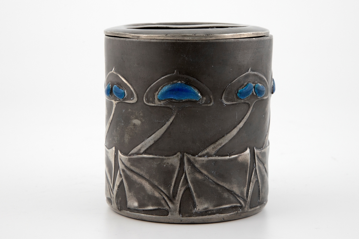 Tobakksdåse med lokk i tinn, dekorert med emalje. Dåsens sylinderformede korpus består av to halvdeler som er loddet sammen. Langs nedre del av korpus er det fremstilt en frise av stiliserte blomstermotiver, hvor blomstenes blader er dekorert med blåfarget opak emalje.