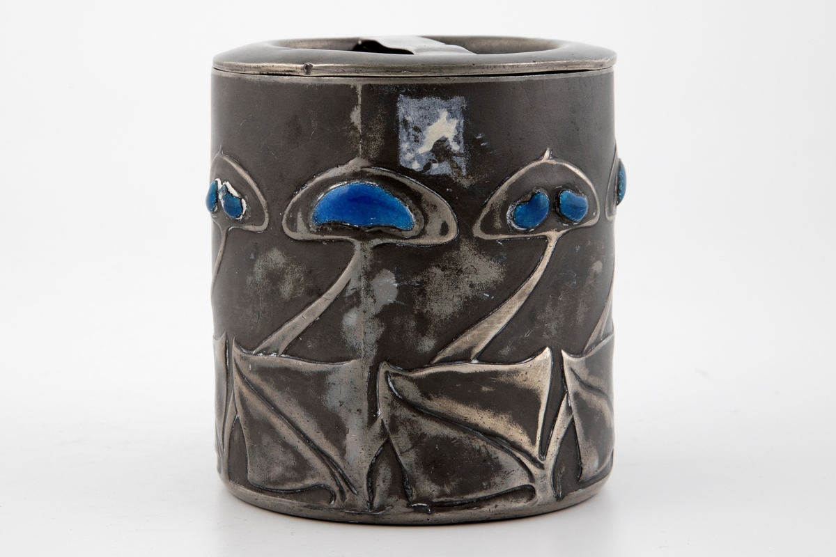 Tobakksdåse med lokk i tinn, dekorert med emalje. Dåsens sylinderformede korpus består av to halvdeler som er loddet sammen. Langs nedre del av korpus er det fremstilt en frise av stiliserte blomstermotiver, hvor blomstenes blader er dekorert med blåfarget opak emalje.