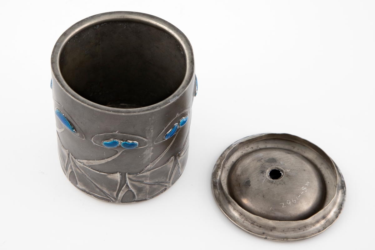 Tobakksdåse med lokk i tinn, dekorert med emalje. Dåsens sylinderformede korpus består av to halvdeler som er loddet sammen. Langs nedre del av korpus er det fremstilt en frise av stiliserte blomstermotiver, hvor blomstenes blader er dekorert med blåfarget opak emalje.