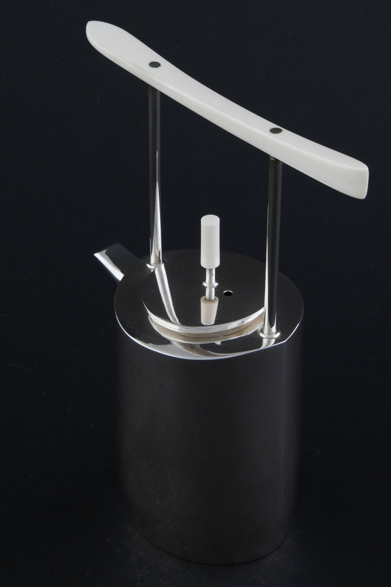 Kaffekanne i sølv. Ovalformet korpus med lett bølgende vegger og liten tut. Fra skulderen reiser det seg to tynne rør som forbinder kannen med et håndtak i elfenben. Sirkulært lokk med sylinderformet lokknapp i elfenben. 